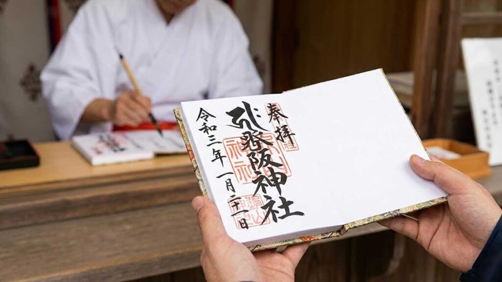 A Shinto priest writing handwritten calligraphy and pressing a red stamp (Goshuin) into a visitor's Goshuin-cho book